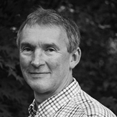 A black and white headshot of Simon Davison, turned to the left and looking at the camera with a lovely smile